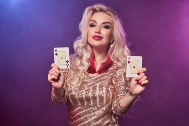 Sarışın, muhteşem bir kadının mükemmel saç stili ve parlak makyajlı, altın sarısı bir elbise giymiş, elinde iskambil kâğıdıyla poz veren ve gülümseyen bir fotoğrafı. Siyah dumanlı arka planda pembe ve mavi arka farlı poker konsepti. Casi.