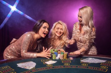Mükemmel saç stilleri ve parlak makyajlı, altın gibi parlak elbiseler giymiş güzel kadınlar kumar masasında durup gülüyorlar. Siyah dumanlı arka planda pembe ve mavi arka farlı poker konsepti. Kumarhane.