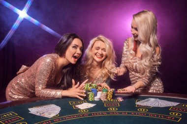 Beautiful females with a perfect hairstyles and bright make-up, dressed in a golden shiny dresses are posing standing at a gambling table and laughing. Poker concept on a black smoke background with pink and blue backlights. Casino. clipart