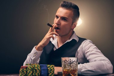 Beyaz gömlekli ve siyah yelekli ciddi bir yetişkin puro içiyor ve kameraya bakıyor. Kumarhanede arka planda siyah arka planda poker oynuyor. Kartlar ve fişler ile riskli kumar eğlencesi.