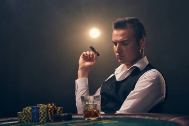 Beyaz gömlekli ve siyah yelekli zengin bir erkek elinde bir puro tutuyor ve aşağıya bakıyor. Kumarhanede arka planda siyah arka planda poker oynuyor. Kartlar ve fişler ile riskli kumar eğlencesi.