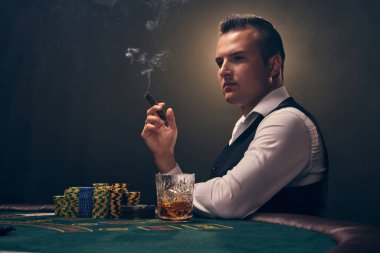Beyaz gömlekli ve siyah yelekli yakışıklı bir yetişkin masada yan yana oturmuş puro içiyor ve başka tarafa bakıyor. Kumarhanede arka planda siyah arka planda poker oynuyor. Kartlar ve çiplerle riskli kumar eğlencesi