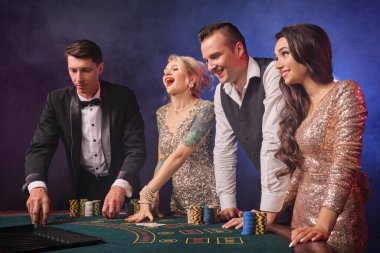 Gazinoda duman içinde poker oynayan neşeli zengin sınıf arkadaşlarının yan fotoğrafları. Gençler büyük bir galibiyet için bahis oynuyorlar. Onlar siyah arka planda kırmızı ve mavi bir arka ışığa karşı masada duruyorlar. Riskli kumar eğlencesi.