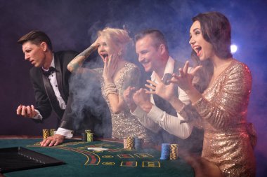 Kumarhanede duman içinde poker oynayan neşeli zengin arkadaşlarının yan fotoğrafları. Gençler büyük bir galibiyet için bahis oynuyorlar. Zaferlerini kutluyorlar. Siyah arka plandaki kırmızı ve mavi ışıklara karşı masada duruyorlar. Riskli bir kumar.
