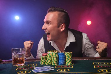 Beyaz gömlekli ve siyah yelekli zarif olgun bir adam, siyah duman arka planında kırmızı ve mavi bir arka fona karşı kumarhanede elinde cipsler ve bir bardak viskiyle poker masasında poz verirken mutlu görünüyor. Riskli kumar eğlencesi.