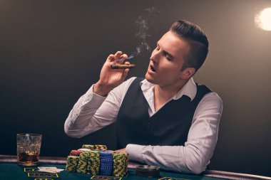 Brunet, beyaz gömlekli ve siyah yelekli olgun bir adam puro içiyor ve ona bakıyor. Kumarhanede arka planda siyah arka planda poker oynuyor. Kartlar ve fişler ile riskli kumar eğlencesi.