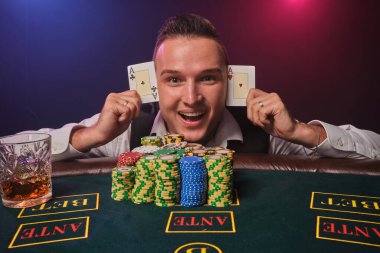 Beyaz gömlekli ve siyah yelekli zarif olgun bir adam elinde iki iskambil kâğıdı tutuyor ve kameraya bakıyor. Poker masasında elinde cipsler ve bir bardak viskiyle poz veriyor. Kumarhanede, siyah üzerine kırmızı ve mavi bir arka farla.