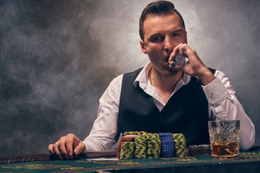 Beyaz gömlek ve siyah yelek giyen yakışıklı olgun bir insan puro içiyor ve kameraya bakıyor. Kumarhanede arka planda siyah dumanla poker oynuyor. Kartlar ve fişler ile riskli kumar eğlencesi.