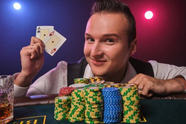 Beyaz gömlekli ve siyah yelekli atletik olgun bir adam elinde iki iskambil kağıdı gösteriyor ve gülümsüyor. Poker masasında üzerinde cipsler ve bir bardak viskiyle poz veriyor. Kumarhanede, siyah arka planda kırmızı ve mavi bir fona karşı. R
