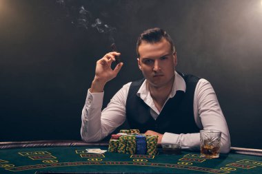 Beyaz gömlekli ve siyah yelekli yakışıklı olgun bir adam masada oturmuş puro içiyor. Kumarhanede arka planda siyah arka planda poker oynuyor. Kartlar ve fişler ile riskli kumar eğlencesi.