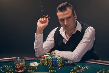 Brunet beyaz gömlekli ve siyah yelekli olgun bir adam puro içiyor ve kameraya bakıyor. Kumarhanede siyah arka planda bir ışık hüzmesiyle poker oynuyor. Kartlar ve fişler ile riskli kumar eğlencesi.
