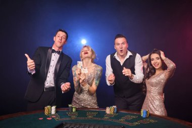 Bir grup zengin sınıf arkadaşı kumarhanede poker oynuyor. Gençler büyük bir galibiyet için bahis oynuyorlar. Masada duran kameraya bakıyorlar. Siyah arka plandaki kırmızı ve mavi arka farlara bakıyorlar. Riskli kumar eğlencesi.