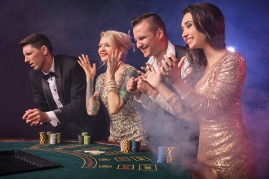 Kumarhanede duman içinde poker oynayan çok mutlu zengin dostların yan fotoğrafları. Gençler büyük bir galibiyet için bahis oynuyorlar. Siyah arka plandaki kırmızı ve mavi arka farlara karşı ellerini çırpıyorlar. Riskli kumar eğlencesi