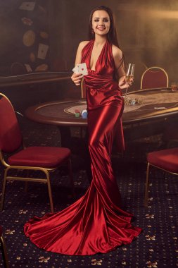 Uzun burgundy atlas elbisesi içinde elinde iki as ve elinde bir bardak şampanyayla genç esmer kız VIP kumarhanesinde poker masasına poz veriyor. Tutku, kumar, cips, alkol, içki, kazanmak, kumar - en iyi eğlence..