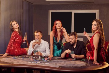 Bir grup genç oyuncu kumarhanede poker masasında poz veriyor ve kameraya bakıyor. Gençler büyük bir galibiyet için bahis oynuyorlar. Para için kumar oynuyorum. Şans oyunları..