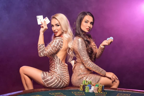 Attractive girls with an excellent hairstyles and professional make-up, dressed in a golden sparkling dresses are posing sitting on a gambling table with some money and chips in their hands and looking at the camera. Poker concept on a black smoke ba