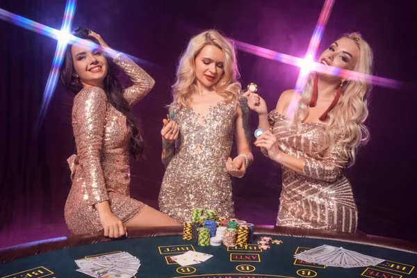 完璧な髪型と明るい化粧をした美しい女性は 金色の光沢のあるドレスを着てギャンブルテーブルに立ってポーズをとっています ピンクと青のバックライトと黒の煙の背景にポーカーコンセプト カジノ — ストック写真