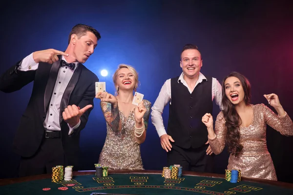 エレガントな豊かな仲間のグループは カジノでポーカーをプレイしています 若者は大きな勝利を待って賭けをしています 黒を背景に赤と青のバックライトを背にテーブルに立っている喜びを感じています リスクの高いギャンブルエンターテイメント — ストック写真
