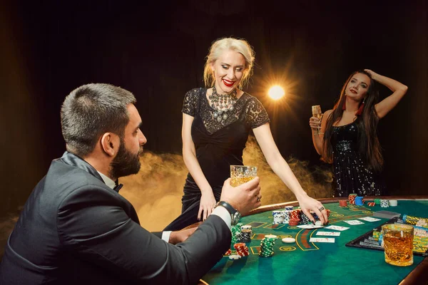 Group of young rich people is playing poker in the casino. Man in business suit and two young women in black dresses. Smoke. Casino. Poker