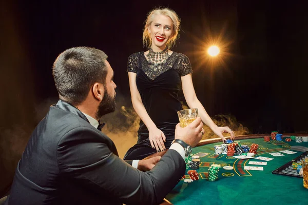 Group of young rich people is playing poker in the casino. Man in business suit and two young women in black dresses. Smoke. Casino. Poker