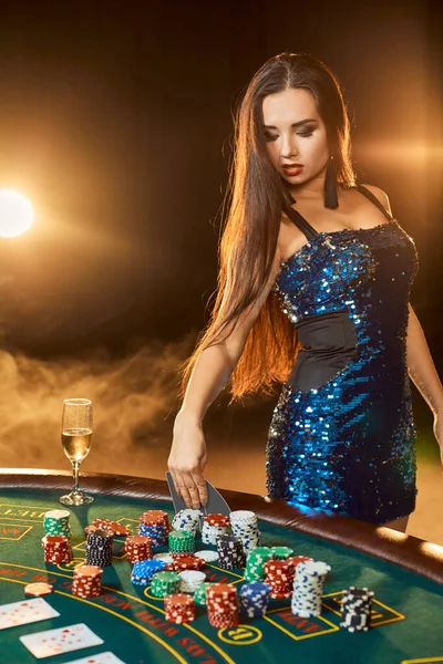 Young beautiful woman in a blue shiny dress poses near poker table in luxury casino. Woman player. Passion, cards, chips, alcohol, dice, gambling, casino - it is as female entertainment. Dangerous fun card game for money. Smoke background.