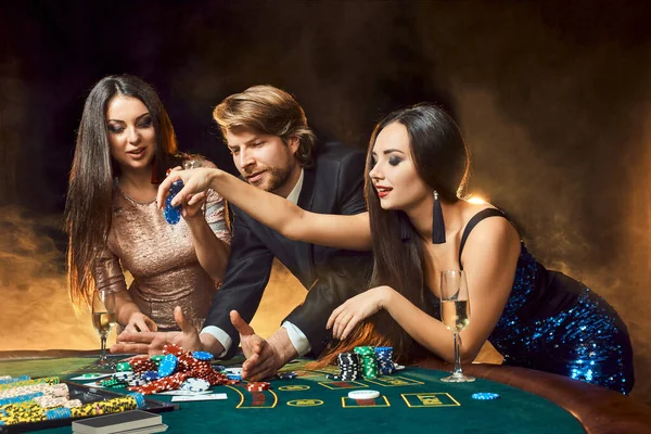 2人の美しい女性と若い男がカジノのポーカーテーブルでプレーし 男とブルネットに焦点を当てます カード チップ アルコール ダイス ギャンブル カジノ それはエンターテイメントです お金のための危険な楽しいカードゲーム — ストック写真