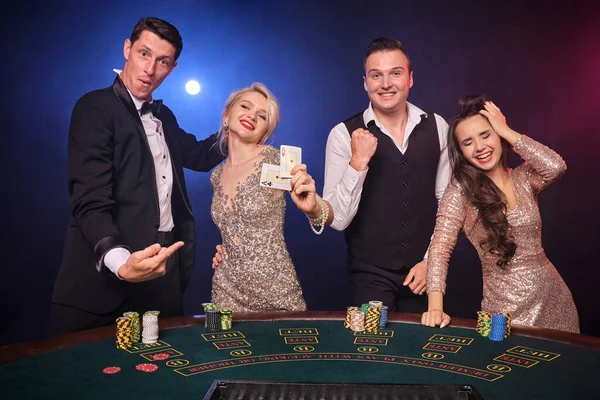スタイリッシュな裕福な仲間のグループは カジノでポーカーをプレイしています 若者は大きな勝利を待って賭けをしています 彼らは黒い背景の赤と青のバックライトに対してテーブルに立って幸せそうに見えます リスクの高いギャンブルエンターテイメント — ストック写真