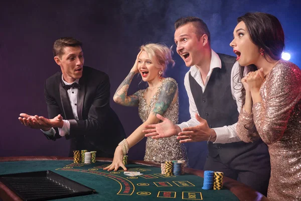 煙の中でカジノでポーカーをプレイ楽しい豊かな友人のサイドショット 若者は大きな勝利を待って賭けをしています 彼らは黒い背景の赤と青のバックライトに対してテーブルに立っているかのように見えます リスクの高いギャンブルエンターテイメント — ストック写真