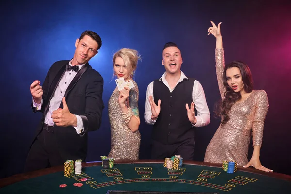 スタイリッシュな裕福な同僚のグループは カジノでポーカーをプレイしています 若者は大きな勝利を待って賭けをしています 彼らは黒を背景に赤と青のバックライトに対してテーブルに立って興奮しています リスクの高いギャンブルエンターテイメント — ストック写真