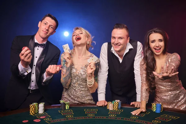 幸せな裕福な同僚のグループがカジノでポーカーをしています 若者は大きな勝利を待って賭けをしています 彼らは黒い背景の赤と青のバックライトに対してテーブルに立っています リスクの高いギャンブルエンターテイメント — ストック写真