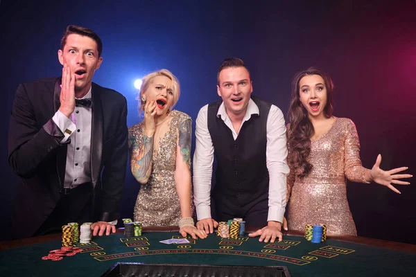 裕福な友人のグループはカジノでポーカーをしています 若者は大きな勝利を待って賭けをしています 彼らは黒い背景の赤と青のバックライトに対してテーブルに立っています リスクの高いギャンブルエンターテイメント — ストック写真