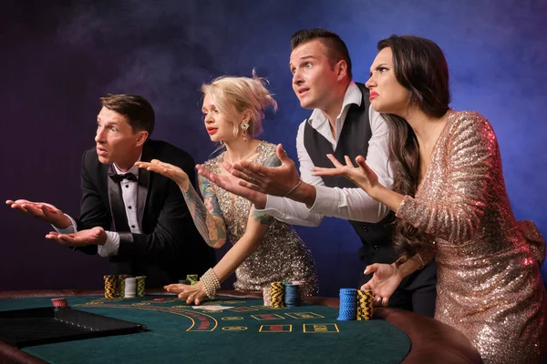 煙の中でカジノでポーカーをプレイ動揺豊かな同僚のサイドショット 若者は大きな勝利を待って賭けをしています 彼らは黒い背景の赤と青のバックライトに対してテーブルに立って不幸な顔をしている リスクの高いギャンブルエンターテイメント — ストック写真