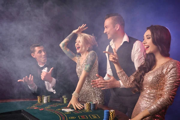 煙の中でカジノでポーカーをプレイする喜びに満ちた豊かなクラスメートのサイドショット 若者は大きな勝利を待って賭けをしています 黒を背景に赤と青のバックライトを背にテーブルに立って笑っている リスクの高いギャンブルエンターテイメント — ストック写真