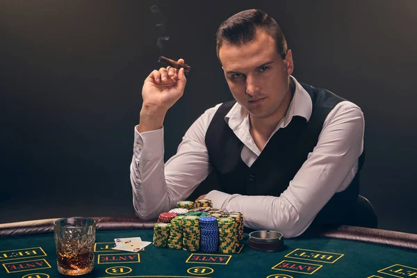 Brunet mature man in a white shirt and black waistcoat is smoking a cigar and looking at the camera. He is playing poker with an excitement at a casino in a ray of backlight on black background. Risky gambling entertainment with cards and chips.