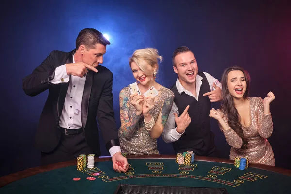 興奮した裕福な友人のグループは カジノでポーカーをしています 若者は大きな勝利を待って賭けをしています 黒を背景に赤と青のバックライトを背にテーブルに立っている喜びを感じています リスクの高いギャンブルエンターテイメント — ストック写真
