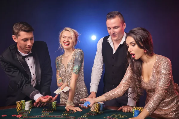 陽気な裕福な同僚のグループはカジノでポーカーをしています 若者は大きな勝利を待って賭けをしています 彼らは黒い背景の赤と青のバックライトに対してテーブルに立っています リスクの高いギャンブルエンターテイメント — ストック写真