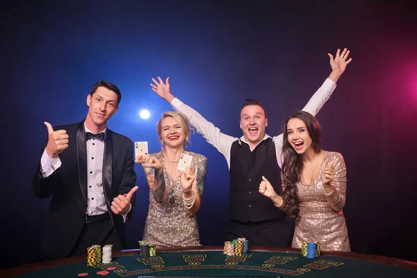 エレガントな豊かな同僚のグループは カジノでポーカーをプレイしています 若者は大きな勝利を待って賭けをしています 黒を背景に赤と青のバックライトを背にテーブルに立って笑っている リスクの高いギャンブルエンターテイメント — ストック写真