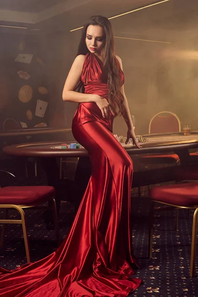 長いバーガンディアトラスのドレスで魅力的なブルネットの乙女が見下ろしており Vipカジノのポーカーテーブルに横に座ってポーズしています カード チップ アルコール飲料 ギャンブル 最高のエンターテイメントです 煙の背景 — ストック写真