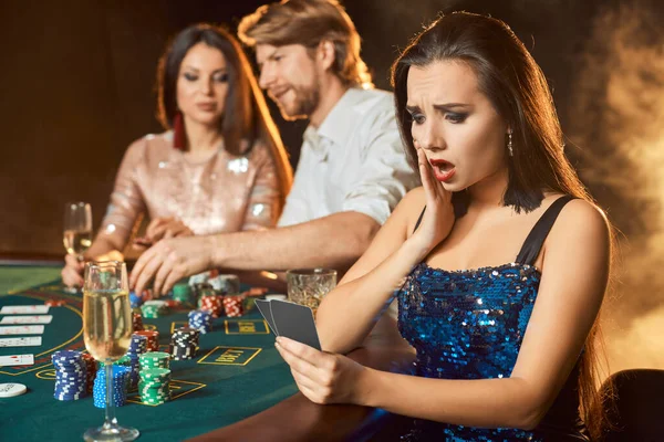 Group of an elegant people playing poker at the gambling house. Focus on a emotional brunette in a blue shiny dress. Passion, cards, chips, alcohol, dice, gambling, casino - it is entertainment. Dangerous fun card game for money. Smoke background.