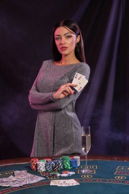 Gri elbiseli muhteşem kadın kumarhanede oyun masasında poz verirken iki kart gösteriyor. Siyah, renkli arka ışıkları olan dumanlı arka plan. Kumar eğlencesi, poker, şampanya. Yakın plan..
