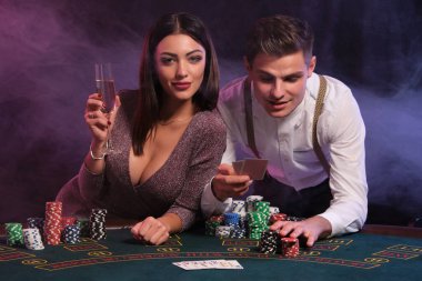 Beyaz tişörtlü adam elinde iki kartla kumarhanede poker oynuyor. Masasında bir sürü fiş var. Kazanmasını seksi elbiseli bir kadınla kutluyor. Siyah, dumanlı arka plan. Kumar oynuyorum. Yakın plan..