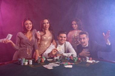Kumarhanede, masada poker oynayan sınıf arkadaşları. Üstünde yığınla fiş, para ve kartlar var. Zaferi şampanyayla kutlamak, gülümsemek. Siyah, dumanlı arka plan, renkli arka ışıklar. Kumar oynuyorum. Yakın plan..