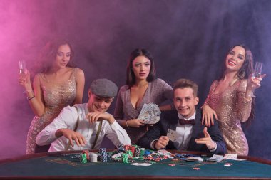 Kumarhanede, masada poker oynayan sınıf arkadaşları. Üstünde yığınla fiş, para ve kartlar var. Kazanmaktan memnun olmak, şampanya içmek, gülümsemek. Siyah, dumanlı arka plan, renkli arka ışıklar. Kumar oynuyorum. Yakın plan..