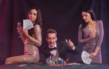 Adam kumarhanede poker oynuyor. Bir sürü fiş, para ve kartla masada oturuyor. Kazanmayı elinde para olan iki bayanla kutluyoruz. Siyah arka plan, renkli arka ışıklar. Kumar, alkol. Yakın plan..