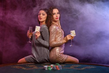 Kumarhanede poker oynayan iki kız ellerinde şampanya ve kağıt bardaklarıyla masada patates kızartması ile oturuyorlar. Siyah, dumanlı arka plan. Yakın plan..