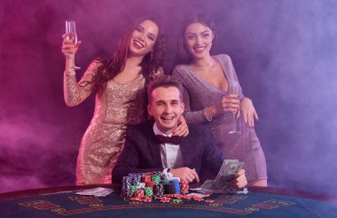 Kumarhanede poker oynayan zarif bir adam ve yanında iki kız var. Üstünde bir yığın fiş ve kartla masada oturmak. Siyah, duman arkaplanı.