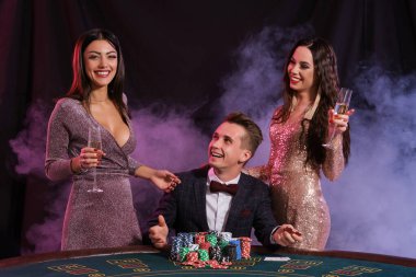 Kumarhanede poker oynayan zarif bir adam ve yanında iki kız var. Üstünde bir yığın fiş ve kartla masada oturmak. Siyah, duman arkaplanı.
