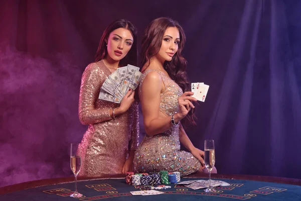 Two ladies in golden dresses showing cards and money, posing at playing table in casino. Black, smoke background, colorful backlights. Gambling entertainment, poker, chips, champagne. Close-up.