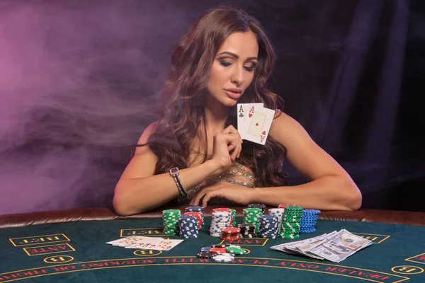 漂亮的少女在赌场打扑克 她拿着两张牌坐在桌旁 上面堆满了薯片和钱 烟熏背景 彩色背光 赌博娱乐 后续行动 — 图库照片
