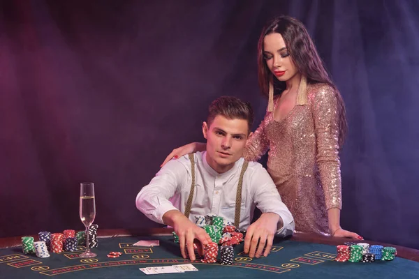 カジノでポーカーをプレイする白いシャツの仲間 それにチップやカードのスタックとテーブルに座って 彼は女性との勝利を祝う 煙の背景 カラフルなバックライト ギャンブルだ — ストック写真